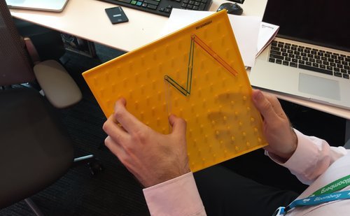 A man has holds a yellow board with pegs. Rubber bands are wrapped around several pegs forming the zig zagging line of a chart.