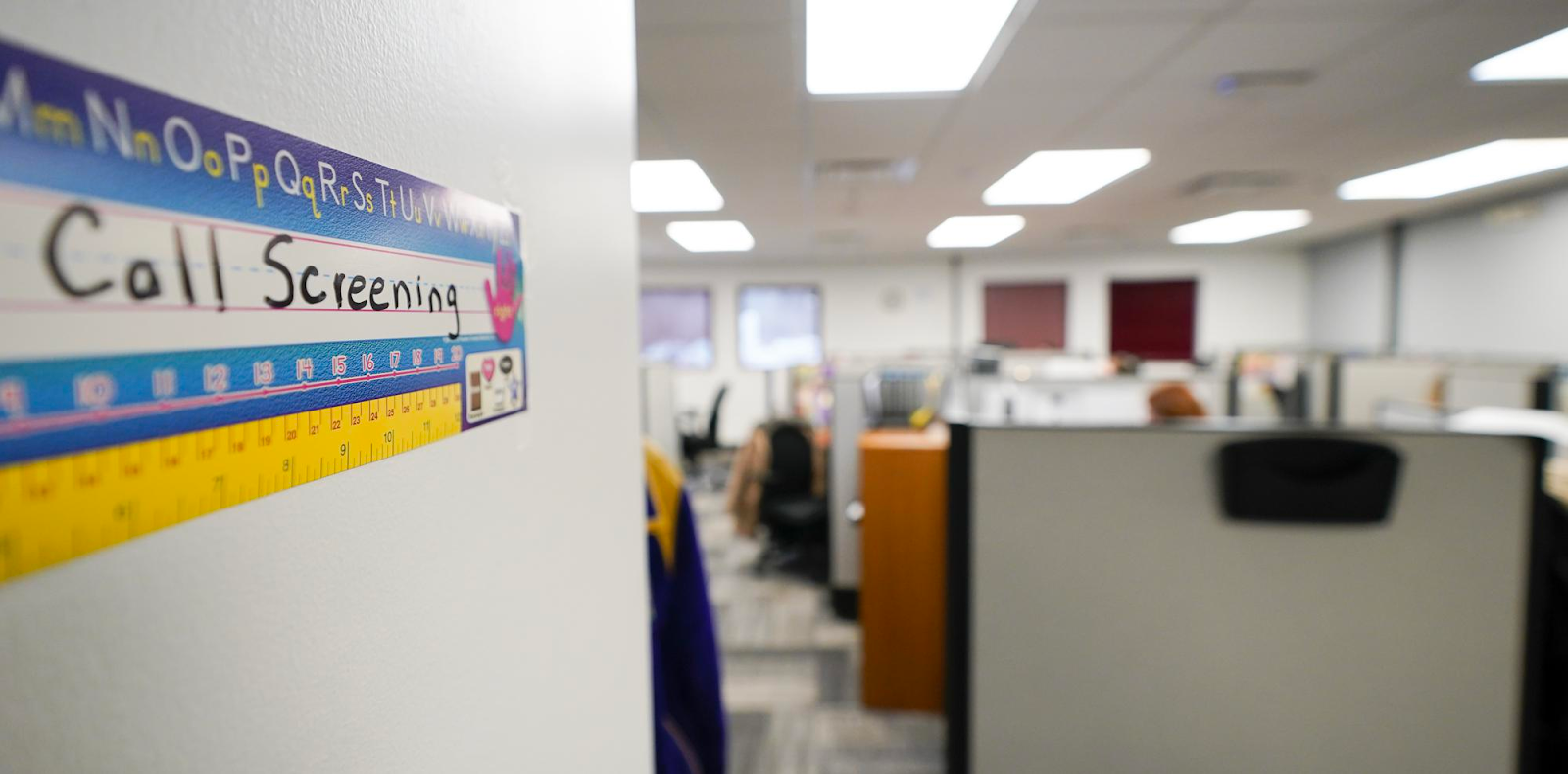 The focus is on a hand written sign that says "Call Screening" on children's lined paper, while an office with cubicles are out of focus in the distance.