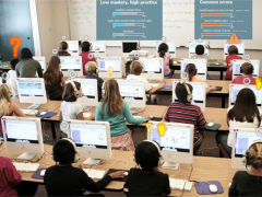 The point of view is from the back of a classroom while students are seated at 4 rows of computers while working on math problems. The teacher can see progress indicator icons above some students, such as a question mark when the student needs help