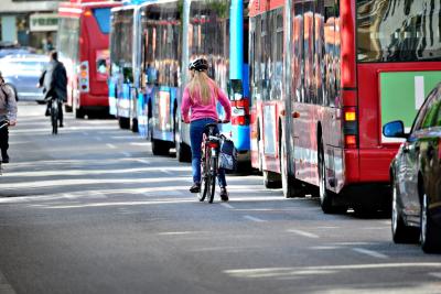 A person with long blond hair and wearing blue jeans, a pink pullover and a helmet rides a bicycle alongside a row of city transit buses of multiple colors in an urban setting.