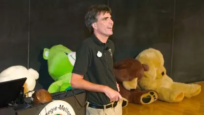 A dark-haired man leans his back against a podium; a chalkboard and many large stuffed animals are behind him.