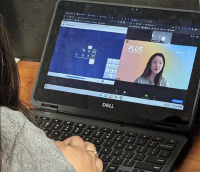 A laptop screen displays two windows: one with a math tutoring problem and another with video of a human tutor.