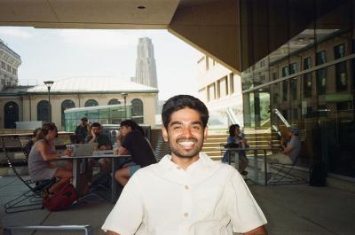 A man sits on a patio, with diners in the background and the Cathedral of Learning in the distance.