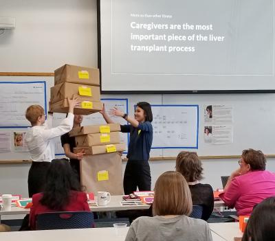 students stack a pile of boxes in the arms of a teammate to illustate the burden of being a caregiver during a capstone presentation