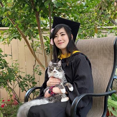 Connie is wearing a black CMU cap and gown. She is sitting outdoors in a chair and holding her cat.