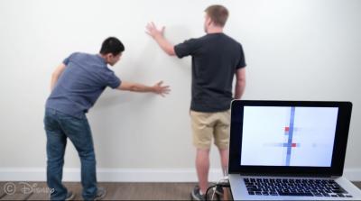 Two men stand in front of a white wall. The locations of where they are touching the wall are visible on a nearby laptop screen.