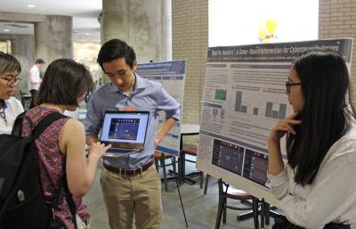 4 students in foreground at a research fair. One student holds laptop open while another student completes a demo. A line of posters on easel down the hall is visible in background