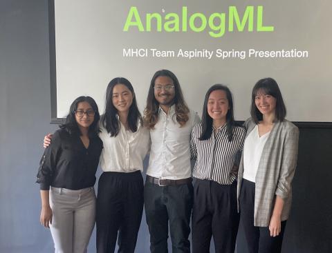 5 team members stand side by side in front of projector screen 