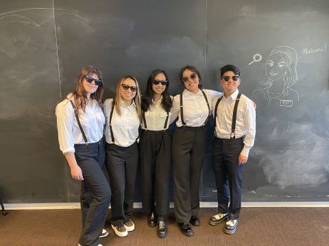 the 5 student team stands side by side, all wearing matching black pants, white shirts, black suspenders, and sunglasses
