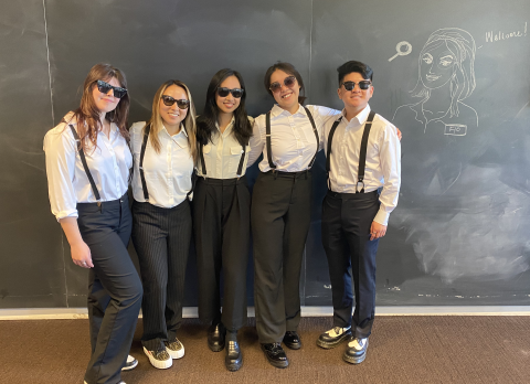 the 5 student team stands side by side, all wearing matching black pants, white shirts, black suspenders, and sunglasses