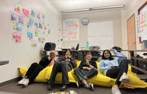 team DPIC chilling on a large yellow beanbag chair, lots of organized post its and affinity diagrams on the walls behind them