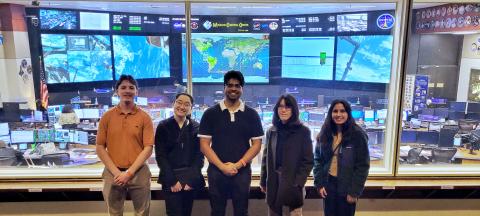 5 students on the NASA Aegis team stand in front of the many screens of Mission Control