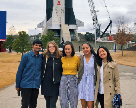 5 members of team NASA stand side by side outdoors in front of a rocket