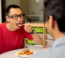 A person sitting at table eating bite of food. There is a small sensor on the side of their glasses