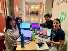 four student team members sit around a table during the hackathon and show their laptop screens while working on Lucid Drums