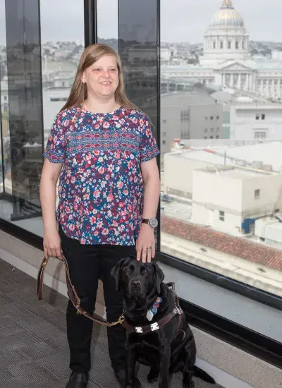 Chancey stands in front of a large window with many buildings in the distance, a black lab service dog sits at her side