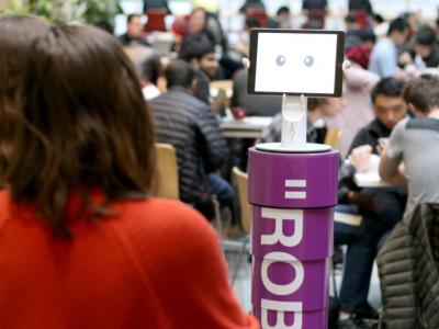 a robot with a purple torso is in the forground of a crowded cafeteria