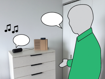 man in a green shirt standing beside DJ bot, which sits on top of a dresser in his home