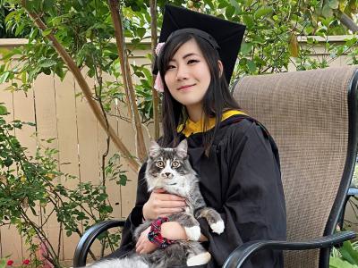 Connie is wearing a black CMU cap and gown. She is sitting outdoors in a chair and holding her cat.
