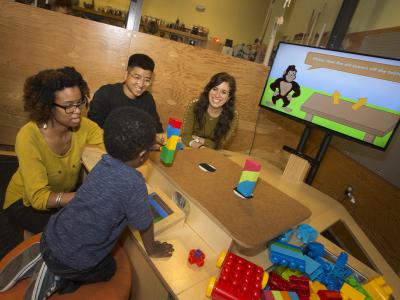3 adults and a child sit at the NoRILLA table and build towers with blocks