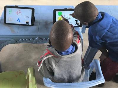 the view of the back two young boys interacting with a touchscreen tablet sitting on a plastic table