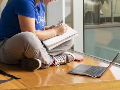 student seated with legs crossed on the floor writing in notebook and laptop open on floor nearby