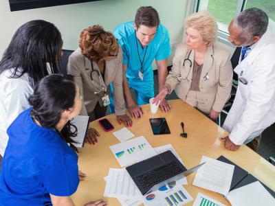 6 doctors and nurses stand around the end of a table looking at charts, graphs and a laptop