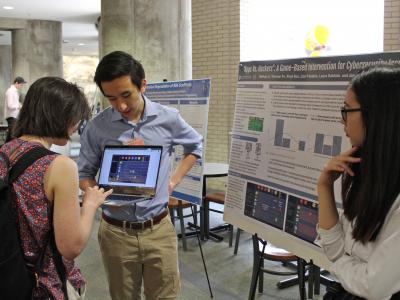 4 students in foreground at a research fair. One student holds laptop open while another student completes a demo. A line of posters on easel down the hall is visible in background
