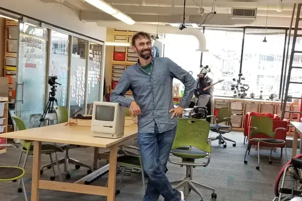 Bigham stands and rests elbow on a small, (vintage?) Apple computer on a table in the lab
