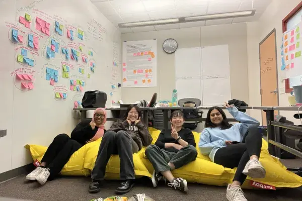 team DPIC chilling on a large yellow beanbag chair, lots of organized post its and affinity diagrams on the walls behind them