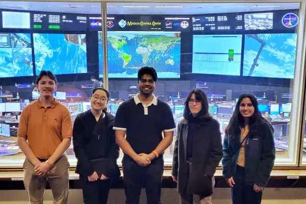 5 students on the NASA Aegis team stand in front of the many screens of Mission Control
