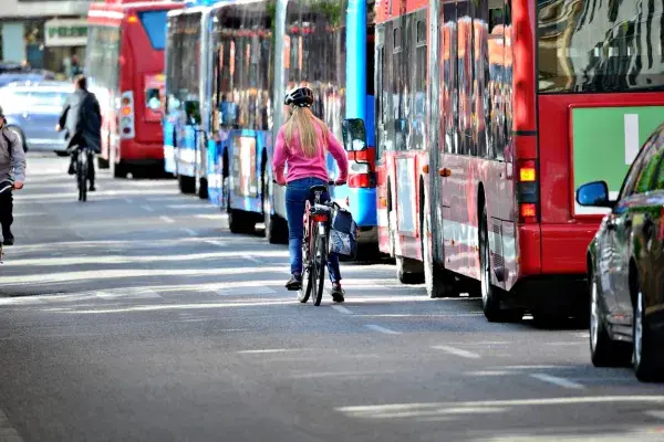 A person with long blond hair and wearing blue jeans, a pink pullover and a helmet rides a bicycle alongside a row of city transit buses of multiple colors in an urban setting.