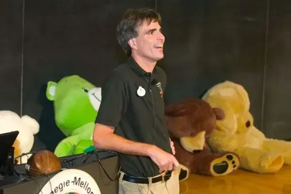 A dark-haired man leans his back against a podium; a chalkboard and many large stuffed animals are behind him.