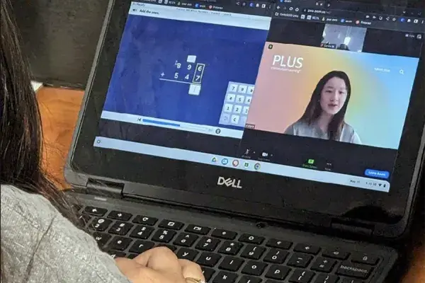 A laptop screen displays two windows: one with a math tutoring problem and another with video of a human tutor.