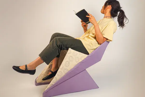 A dark-haired woman with a ponytail holds a book while she reclines on a purple chair.