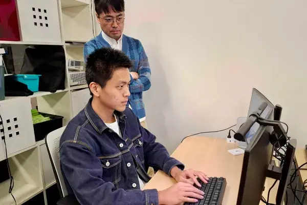 A man works at a desktop computer while another man looks over his shoulder, arms crossed.