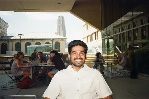 A man sits on a patio, with diners in the background and the Cathedral of Learning in the distance.