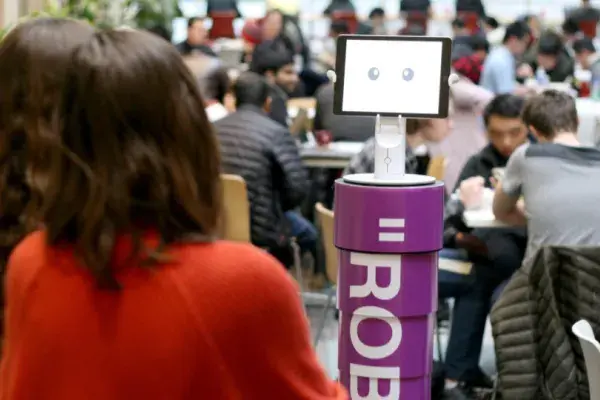 a robot with a purple torso is in the forground of a crowded cafeteria
