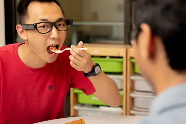 A person sitting at table eating bite of food. There is a small sensor on the side of their glasses