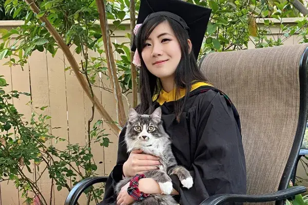 Connie is wearing a black CMU cap and gown. She is sitting outdoors in a chair and holding her cat.