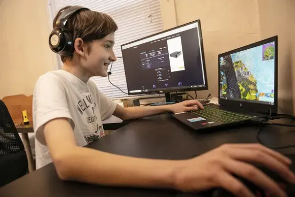 young student smiles while sitting at desk with laptop and extra screen and playing a game online