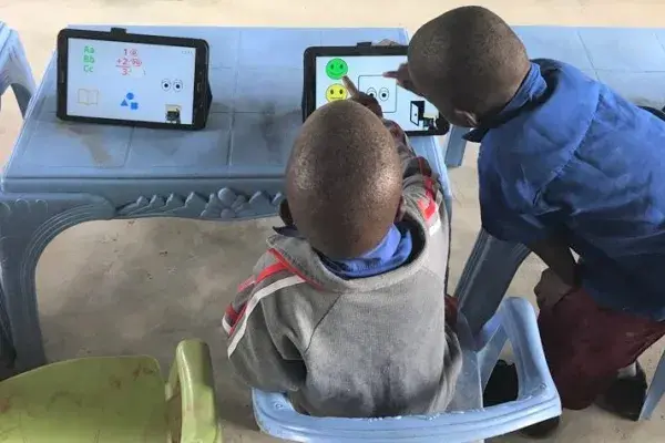 the view of the back two young boys interacting with a touchscreen tablet sitting on a plastic table