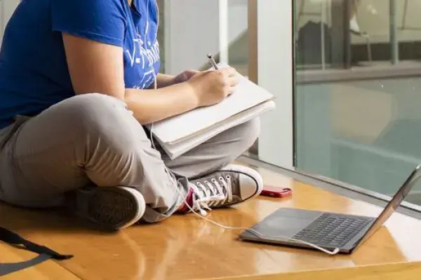 student seated with legs crossed on the floor writing in notebook and laptop open on floor nearby 