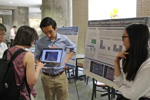 4 students in foreground at a research fair. One student holds laptop open while another student completes a demo. A line of posters on easel down the hall is visible in background